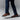 A person is captured mid-step on a neutral background wearing dark blue pants and Anthony Veers Camden Tassel Loafers in Amber Cognac Tan, featuring a premium Italian cowhide and polished hand-burnished patina.