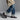 A person wearing cuffed blue jeans, white socks, and Anthony Veer Camden Tassel Loafer in Café Noir Brown with Goodyear welted Italian cowhide leather, plus a beige coat, walks against a plain gray background.