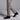 A person wearing light beige dress pants and Anthony Veer Arden Chukka Boot in Velvet Noir Black Suede is stepping forward on a plain grey background. The image, cropped to show just the legs and feet, highlights the stylish suede boots.